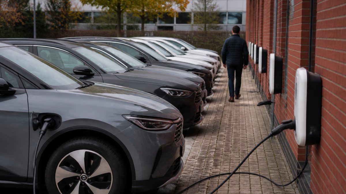 A row of electric vehicles parked in a staff car park with charging cables connected to wall-mounted charge points, a driver walking away in the background