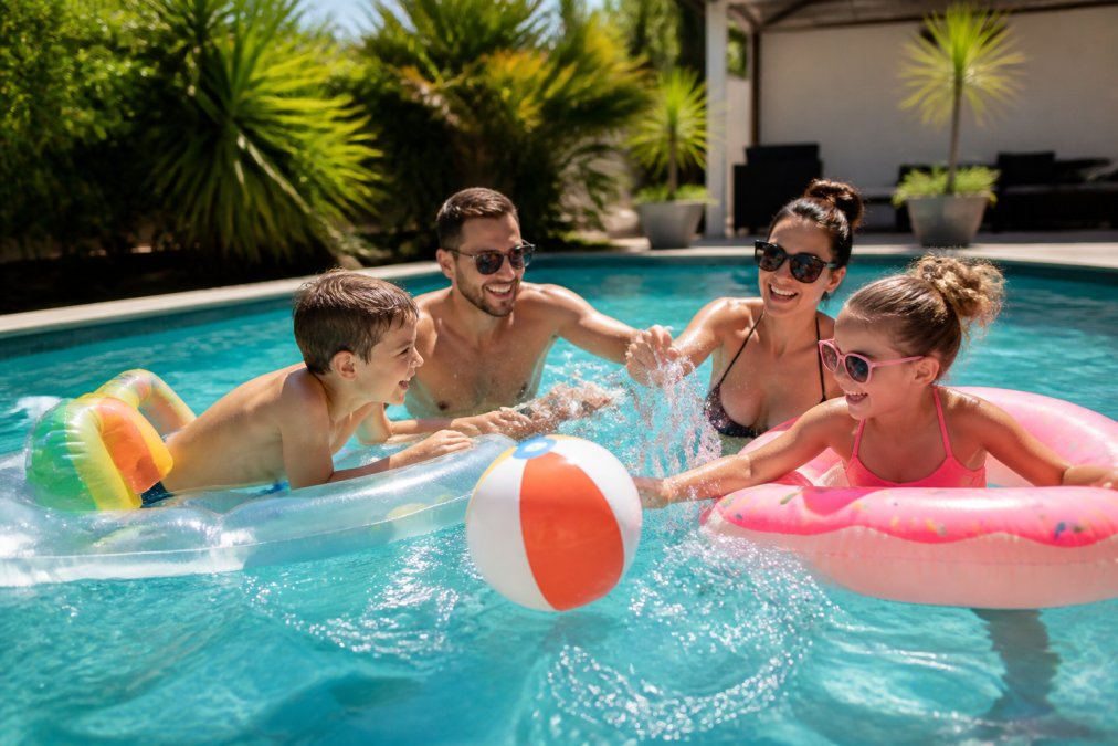 Family of four enjoying various inflatable pool toys and games together in a bright, sunny backyard swimming pool setting, with children laughing and adults splashing