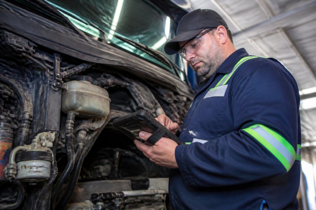 Professional technician performing vehicle inspection in well-lit service bay, using digital tablet to document findings while examining truck engine compartment under bright overhead lighting