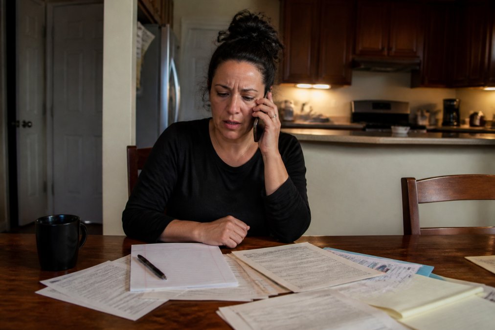 A concerned family member sitting at a kitchen table, speaking on the phone with paperwork and a notepad spread in front of them, warm indoor lighting suggesting a late-night call for help