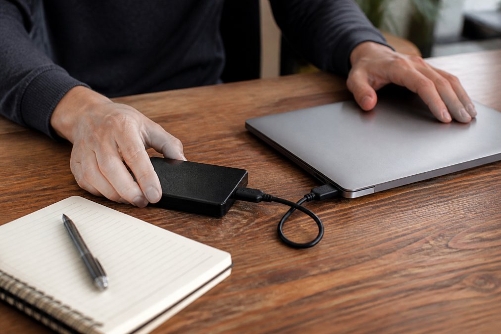 Person backing up laptop files to an external hard drive with a notepad listing important information nearby on a desk