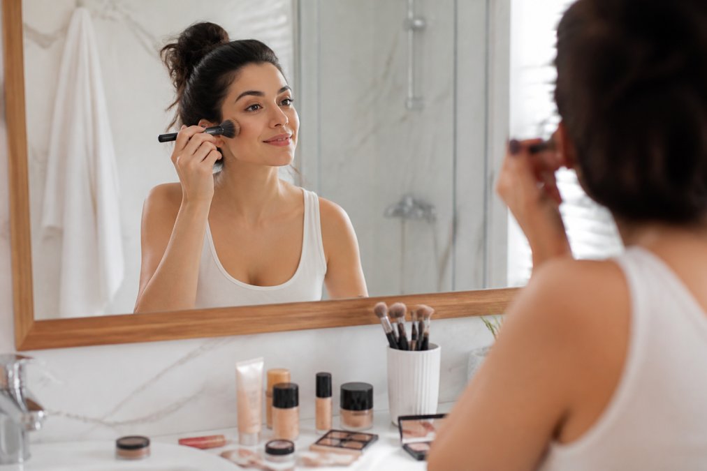 Woman in bright bathroom mirror demonstrating quick makeup application techniques with cosmetic products and brushes visible on counter