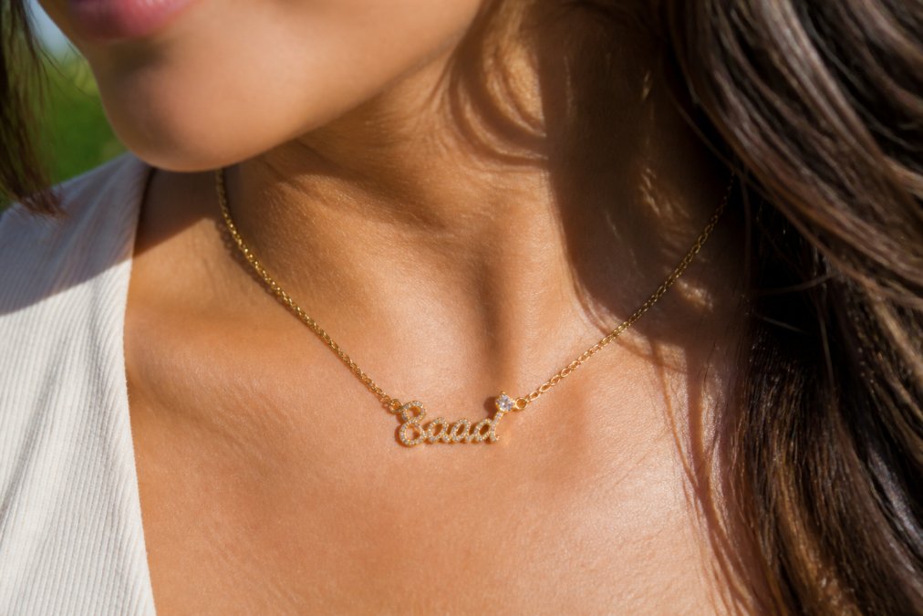 Woman wearing delicate personalized name necklace with cubic zirconia crystals catching natural sunlight, close-up of pendant detail showing clear engraving and sparkle