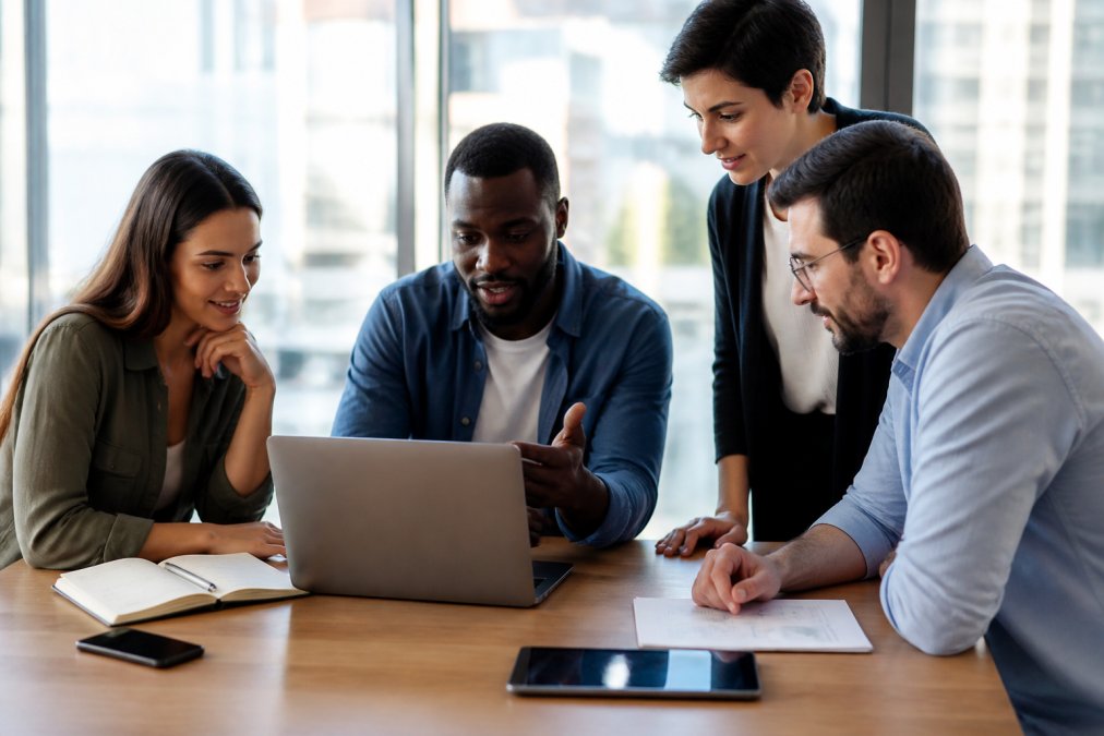 A professional team collaborating around a laptop in a bright, modern office, reviewing a project dashboard with visible task lists on the screen