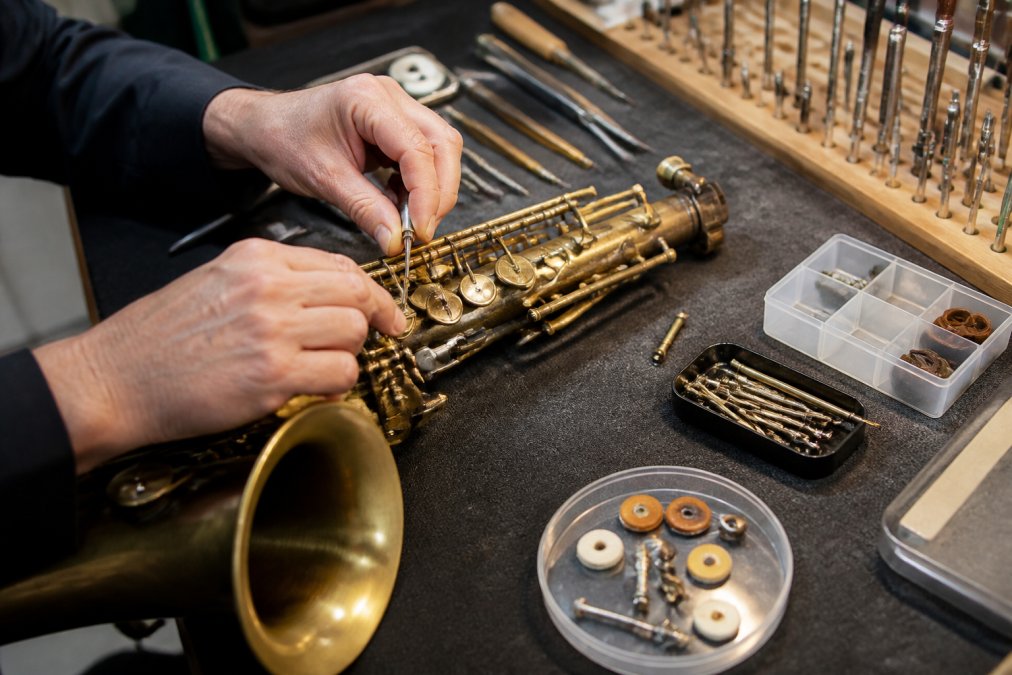 Professional woodwind repair technician's hands working on a saxophone at a repair bench, with precision tools, replacement pads, and metal springs visible under bright workshop lighting