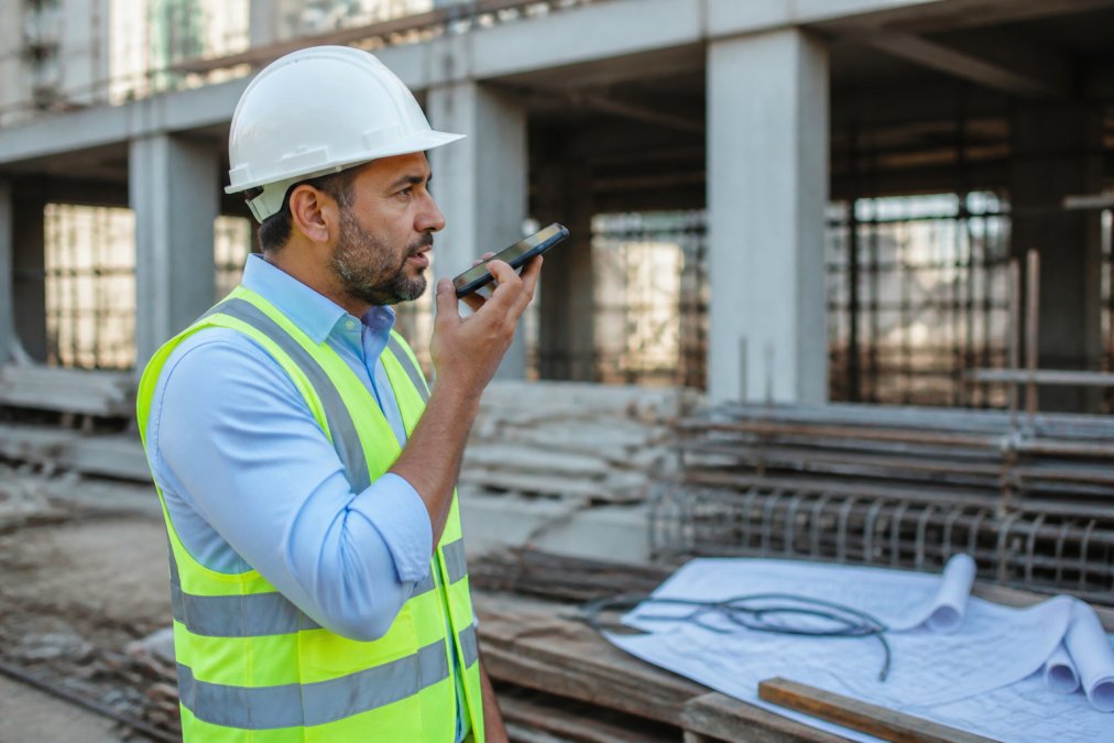Field inspector holding an Android phone to their mouth, dictating notes on a construction site with building materials and blueprints visible in the background, natural daylight