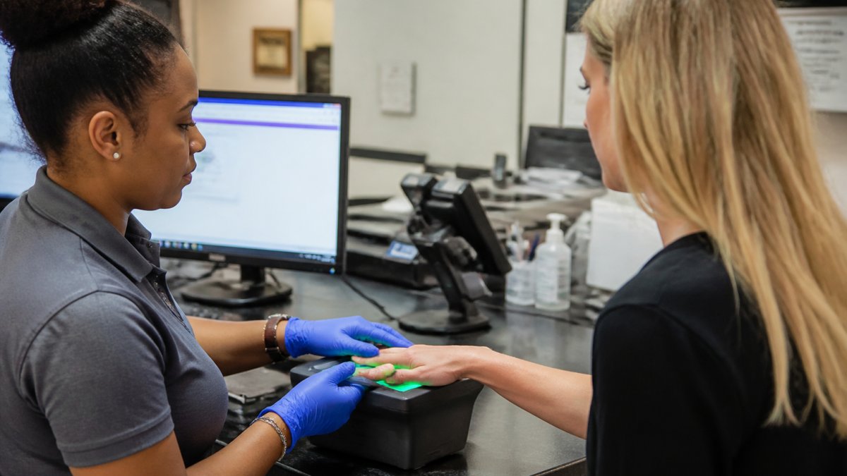 A person having digital fingerprints captured at a professional service counter while a staff member assists, with a computer screen showing a submission portal in the background