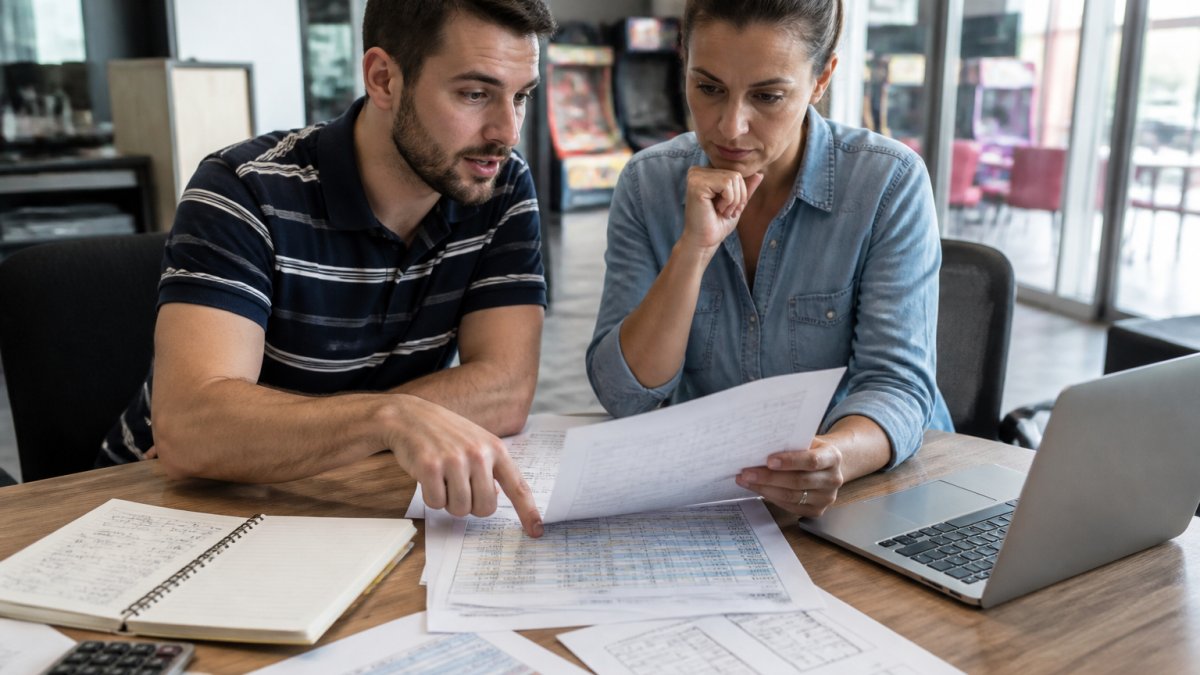 A consultant and a family entertainment center owner reviewing a printed budget and site plans across a table in a bright office, with a laptop showing financial projections nearby