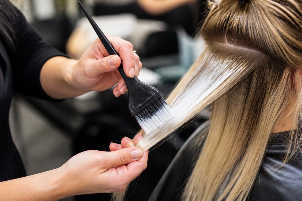 Professional colorist's hands demonstrating feathered brush technique on blonde hair sections, showing the brush held at a 45-degree angle while applying lightener with a soft sweeping motion