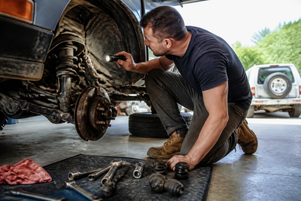 A DIY enthusiast crouching beside the front wheel arch of a lifted car, using a torch to inspect the underside suspension components and steering linkage, in a home garage under natural daylight from an open roller door