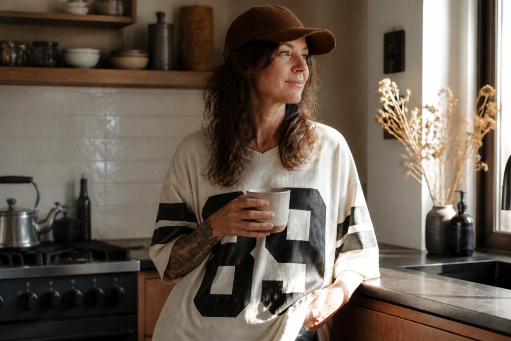 A woman in her forties wearing a corduroy cap and jersey, holding coffee as feel-good gear in a sunny kitchen.
