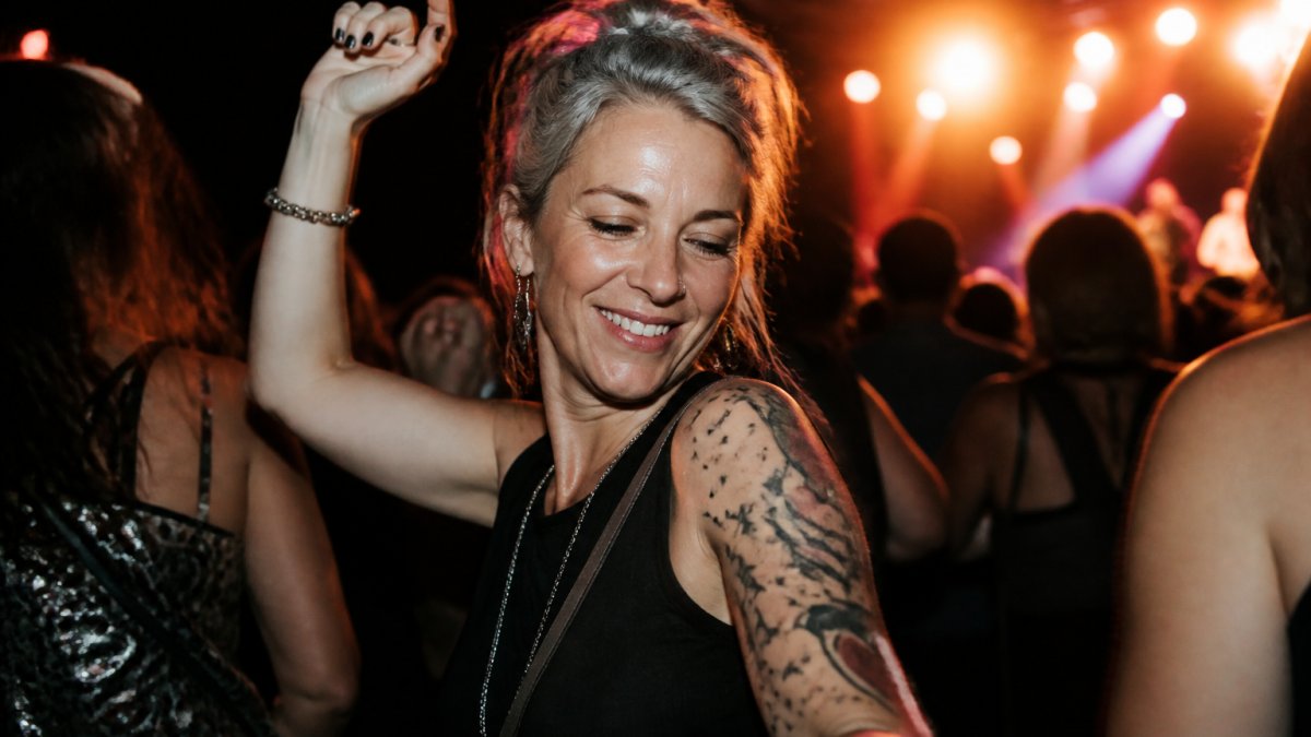 Woman in her forties smiling and dancing with friends at a vibrant live music concert under warm stage lighting.