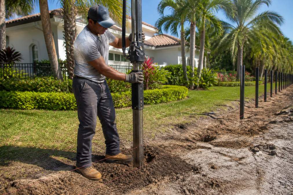 A professional fence installer driving a steel post into sandy, dark South Florida soil beside a large residential property with mature palms and dense tropical landscaping in the background, midday sunlight casting short shadows