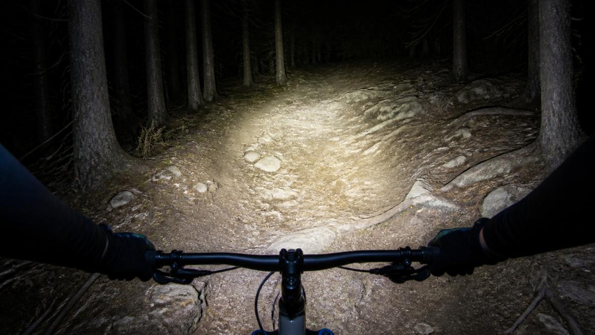 A cyclist riding a mountain bike at night on a dark trail, with a bright LED headlight casting a wide beam of light across the path ahead, illuminating rocks and tree roots