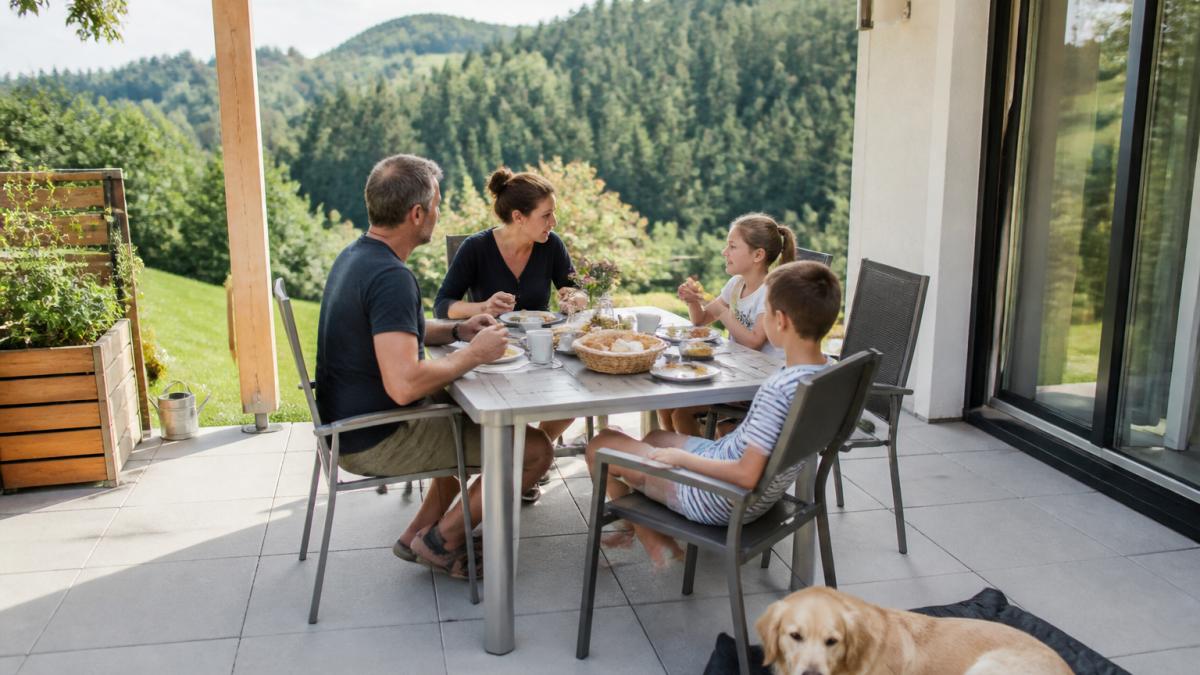 A family with two children is relaxing on the patio of a vacation rental, with the green hills and forests of the Waldviertel in the background; next to them, a dog is lying on a blanket