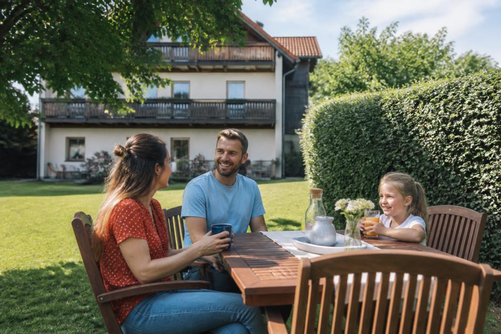 Ein sonniger privater Garten mit einem Holztisch und Stühlen, umgeben von grünen Hecken und Bäumen, ein Ferienwohnungsgebäude im Hintergrund