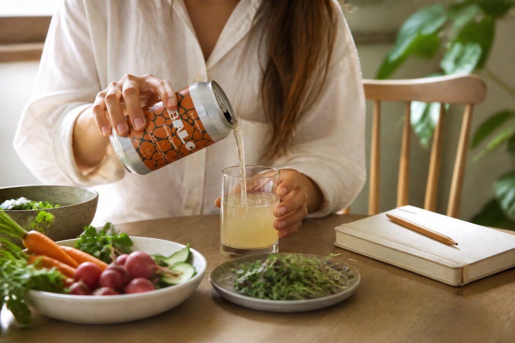 Person pouring fermented vegetable drink into a glass at a dining table with fresh vegetables and wellness journal nearby, natural morning light