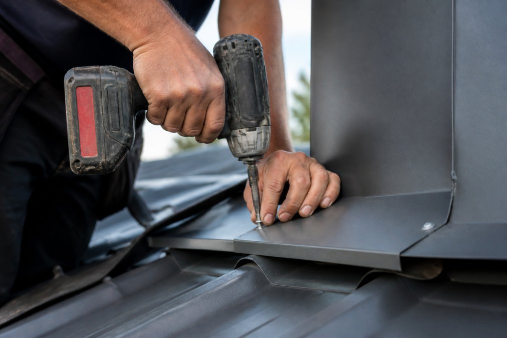 Nærbilde av hendene til en håndverker som monterer en selvborende skrue i en tynn metallplate på et tak, med drill og beslag synlig i bakgrunnen