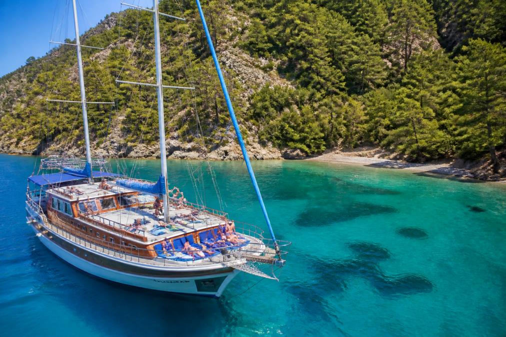Ahşap bir gulet teknesi masmavi turkuaz sularda süzülüyor, güvertede güneşlenen ve manzarayı izleyen turistler, arka planda yemyeşil çam ormanlarıyla kaplı dik yamaçlar ve küçük bir koy
