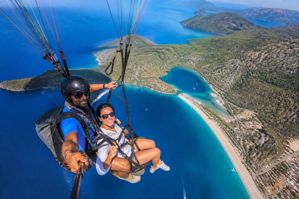 Babadağ'ın zirvesinden havalanan tandem yamaç paraşütü pilotu ve yolcusu, arka planda Ölüdeniz'in turkuaz lagünü ve Belcekız Plajı'nın geniş açıyla görüldüğü güneşli bir an