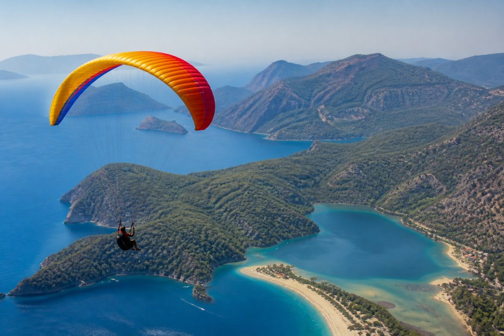 Yamaç paraşütü sporcusu, renkli paraşütüyle Ölüdeniz'in turkuaz sularının üzerinde süzülürken, arka planda Fethiye'nin dağlık coğrafyası ve ormanlık alanlar görülüyor