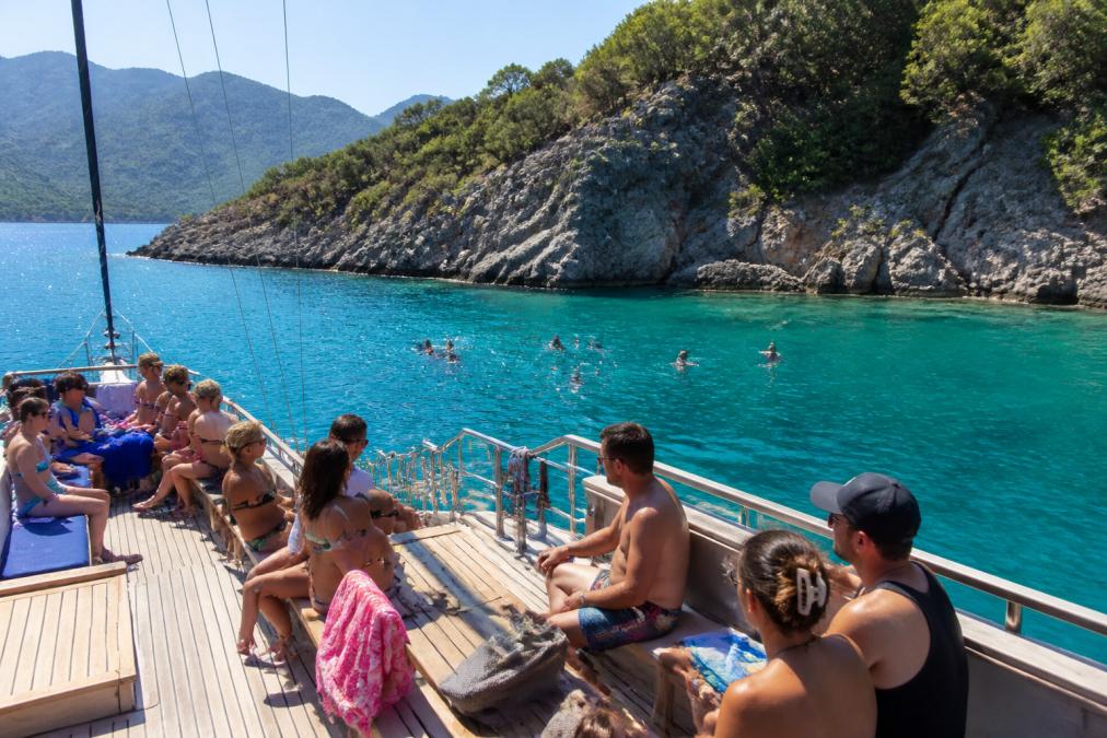 Tekne güvertesinde turist grubu, güneşli hava altında oturmuş, turkuaz denizde yüzen insanlar ve kayalık koylar arka planda, Fethiye'nin dağlık sahili görülüyor