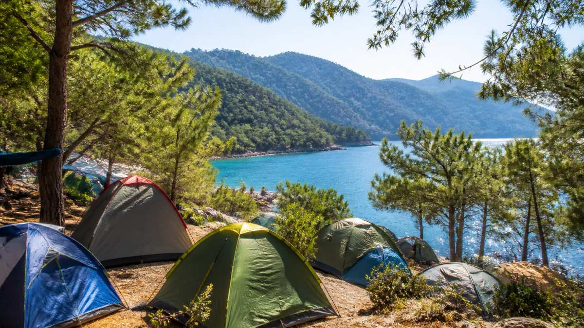 Fethiye'de doğa ortasında kurulan çadırlarla dolu bir kamp alanı, arka planda turkuaz deniz ve yeşil tepeler, sabah güneş ışığında