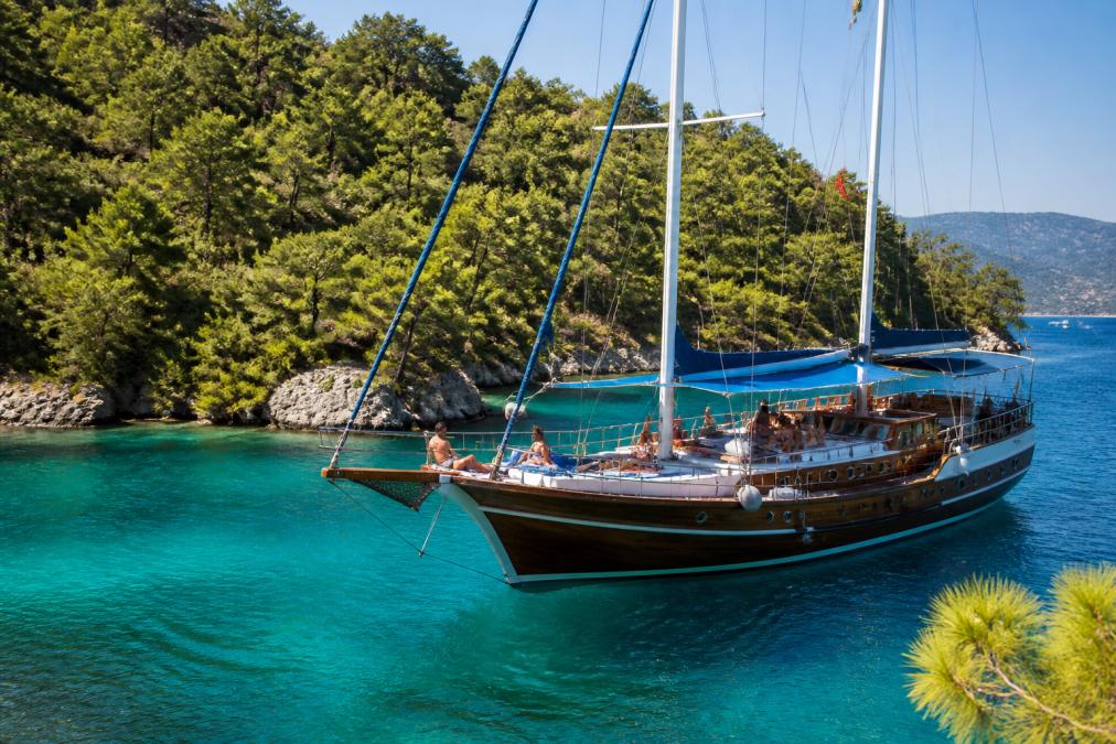 Turkuaz bir koyda demirlemiş ahşap gulet, güvertede güneşlenen ve sohbet eden tatilciler, arka planda çam ormanıyla kaplı yamaçlar