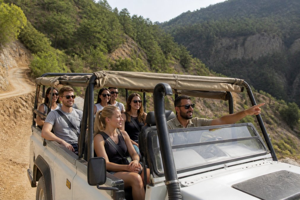 Bir jeep safari aracının içinde emniyet kemeri takan, gülümseyen bir grup turist. Araç, Fethiye'nin ormanlık ve dağlık bir arazisinde toprak yolda duruyor. Rehber ön tarafta eliyle yönü gösteriyor.