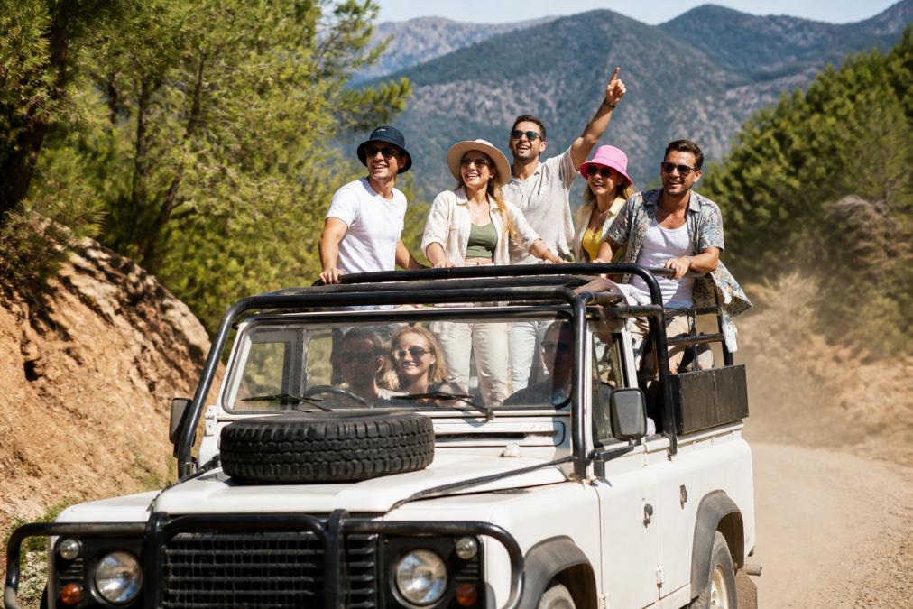Bir safari aracının üzerinde rahat ve açık renkli kıyafetler giymiş gülümseyen bir grup turist, Fethiye'nin ormanlık ve dağlık arazisinde tozlu bir patikada ilerlerken görülüyor.