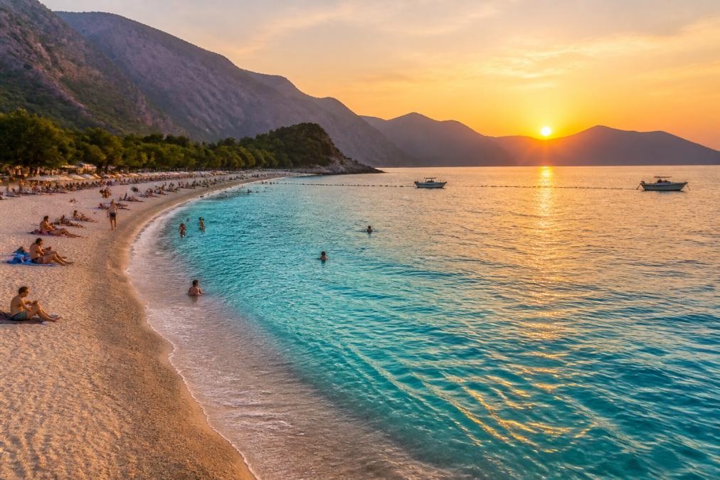Ölüdeniz Plajı'nda kristal berrak turkuaz su, güneşlenen insanlar, kumlu sahil, arka planda dağlar, gün batımı saati