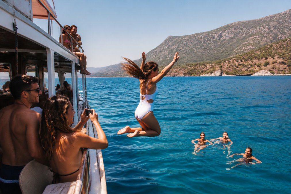Turist grubu tekne turunda denizde eğlenirken, mavi sular ve arkada dağlık Fethiye kıyısı görülüyor, güneşli bir gün