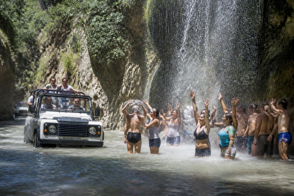 Üstü açık Land Rover cipte, Gizlikent Şelalesi'nin yanında suya giren turist grubu, şelalenin arka planında, güneş ışığında parlayan suların heyecan verici anı