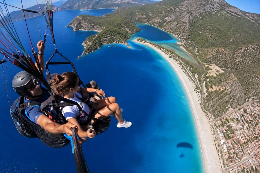 Tandem yamaç paraşütü uçuşu sırasında, Ölüdeniz'in turkuaz denizi, Mavi Lagün ve beyaz kumlu plajlarının kuş bakışı görünümü, paraşütün gölgesi su yüzeyinde