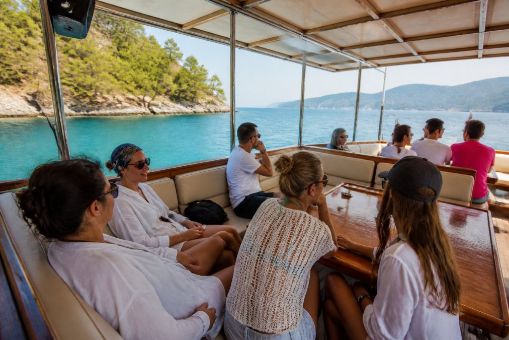 A group of relaxed tourists sitting on the shaded mid-deck of a wooden boat cruising calm turquoise water near pine-covered islands, one person looking out at the horizon with a calm expression