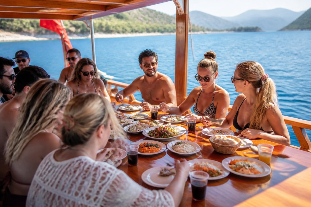 Teknede servis edilen meze tabakları, soğuk içecekler ve öğle yemeği masası, turist grubu güneşli güvertede yemek yiyerek mavi deniz manzarasını izliyor