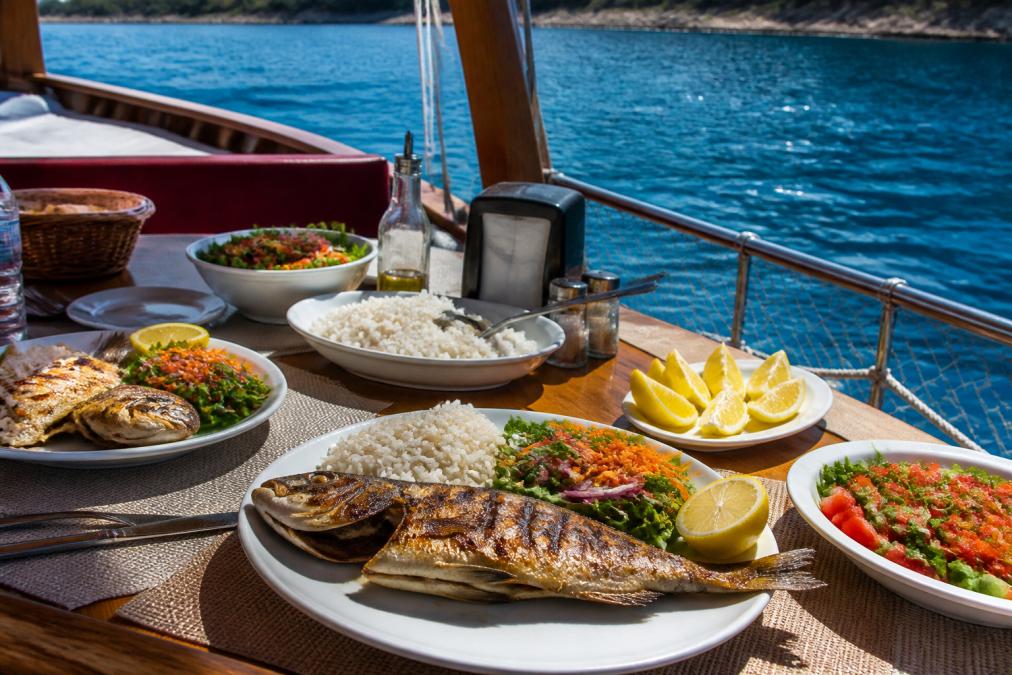 Güneşli bir günde tekne güvertesinde kurulmuş öğle yemeği sofrası; ızgara balık, pilav, taze salata ve limon dilimleriyle dolu tabaklar, arka planda turkuaz deniz
