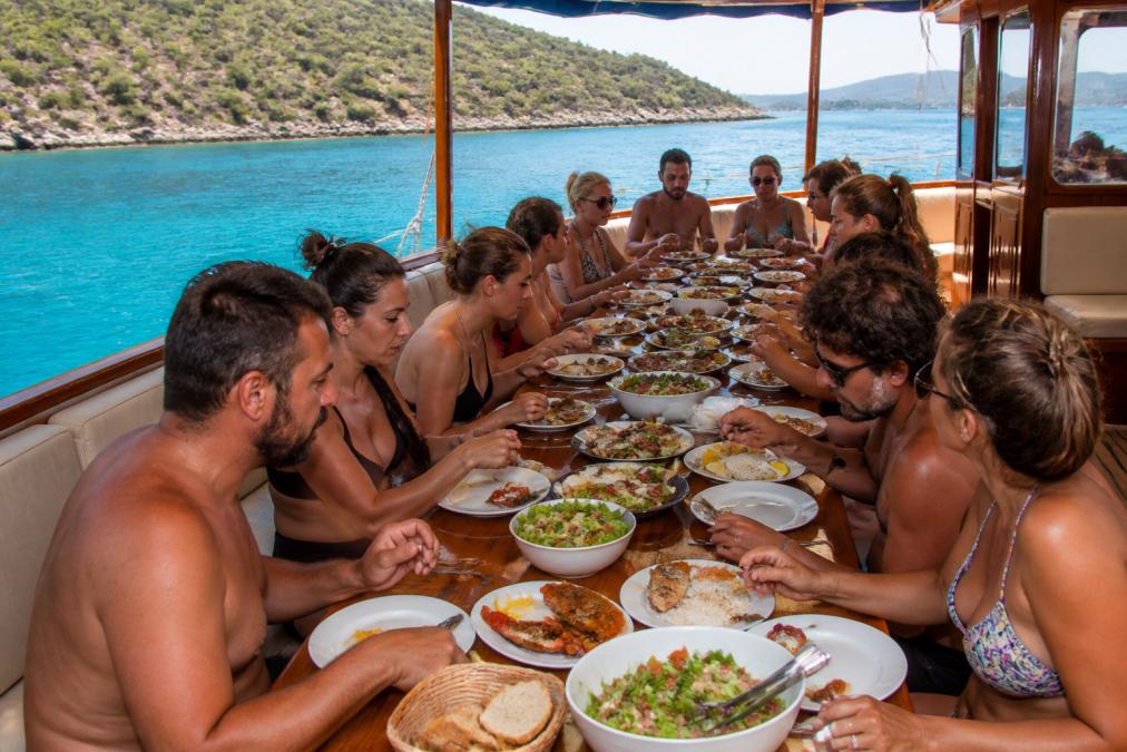 Güneşli bir günde Fethiye koylarında demirlemiş bir teknede uzun bir masada oturan gezginler, taze deniz ürünleri ve mezeleri paylaşıyor; arka planda turkuaz deniz ve çam kaplı yamaçlar