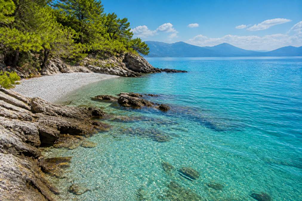 Turkuaz deniz suyu, beyaz kumlu plaj ve kayalık sahil formasyonuyla çevrili sakin bir Fethiye koyu, güneşli bir günde, ufukta dağlar görünen manzara