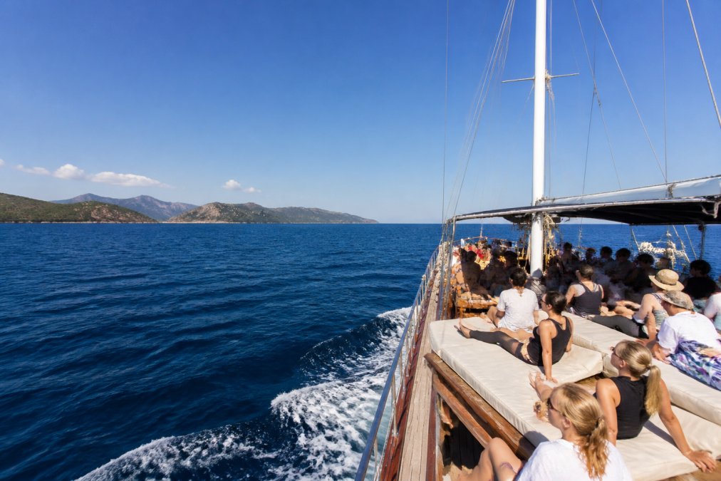 Turist grubuyla dolu tekne, açık denizde yelken açmış, arka planda Fethiye'nin adalarının silueti görünen sahne, güneşli hava