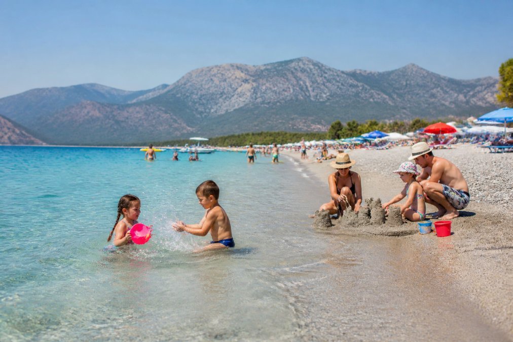 Ölüdeniz plajında sığ, dalga az deniz suyunda oynayan çocuklar, kumda oyun oynayan aileler, berrak mavi su ve dağlar arka planda