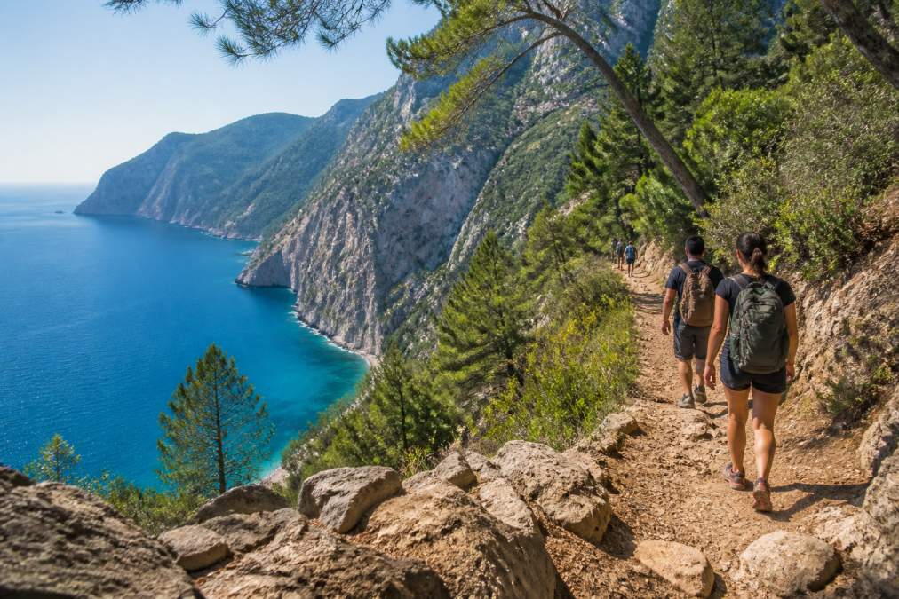 Hikers walking along a cliffside trail with the turquoise sea and steep Butterfly Valley cliffs in the background, warm afternoon sunlight, Mediterranean pine trees framing the path