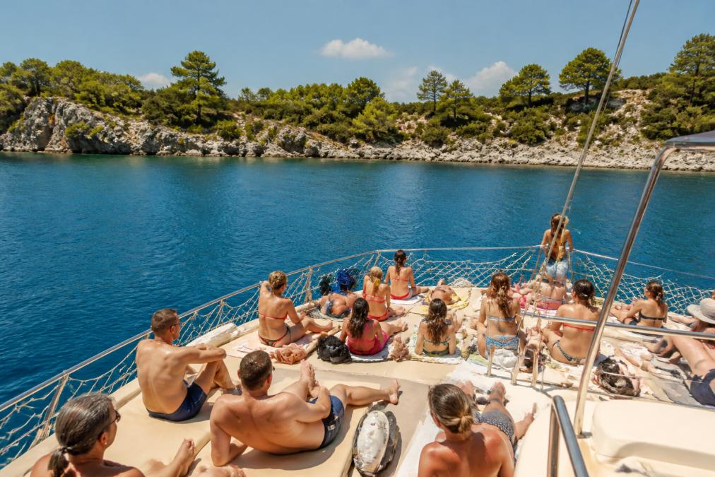 Geniş bir teknenin güvertesinde turist grupları güneşleniyor, arka planda Fethiye'nin berrak mavi suları ve kayalık kıyıları görülüyor