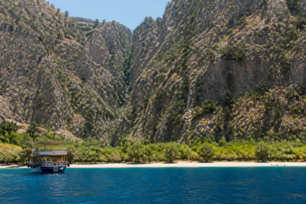 Kelebekler Vadisi'nin yeşil ormanları ve dar kanyonundan akan su, tekne ile yaklaşan ziyaretçiler, dağ arka planında güneşli bir gün