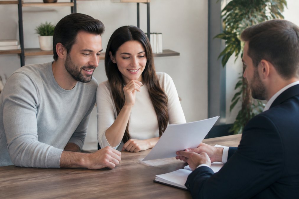 First-time homebuyer couple reviewing mortgage documents and loan comparison materials with a loan officer at a wooden desk in a bright, professional office setting