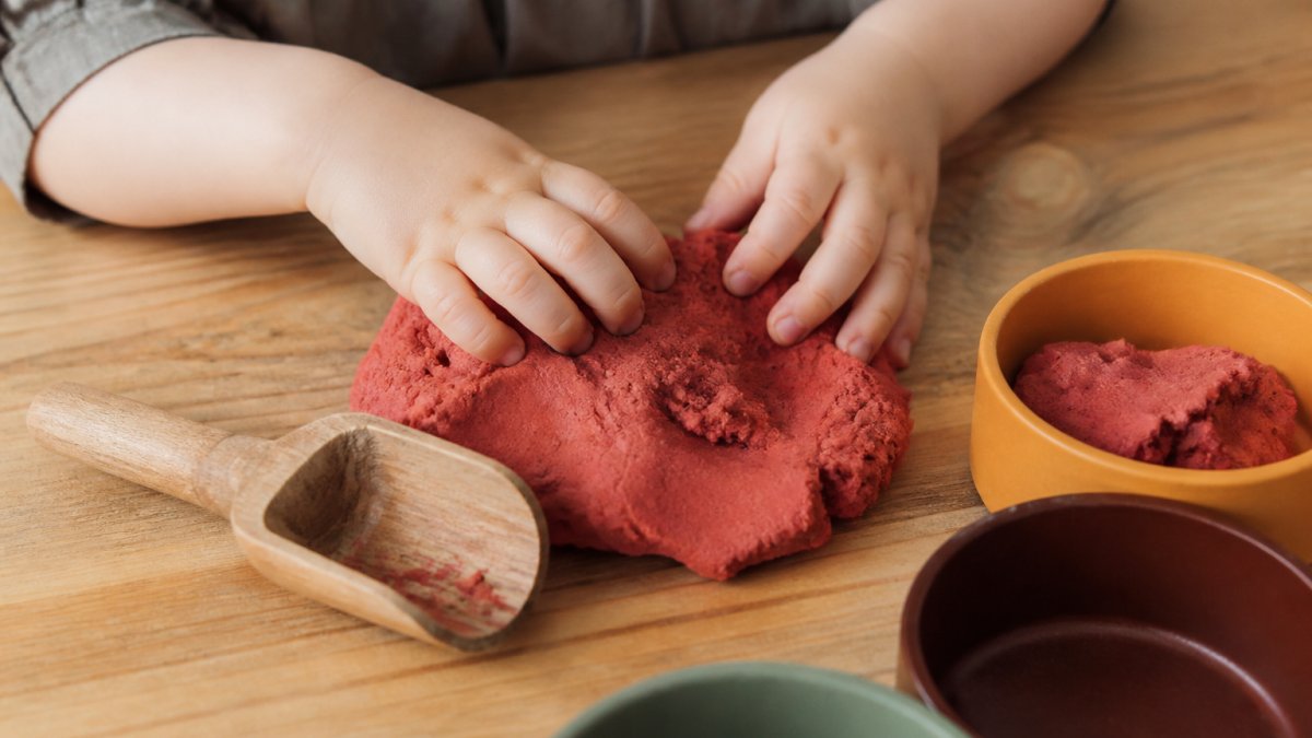 Close-up van peuterhanden die rode natuurlijke speelklei kneden op een houten tafel, met een houten speelschepje en kleurrijke bakjes ernaast