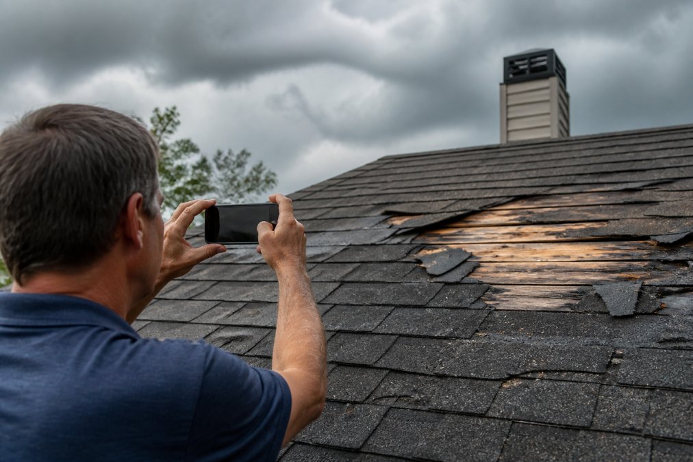 Homeowner photographing roof damage with smartphone, capturing close-up view of torn shingles and exposed wood, with storm clouds in background