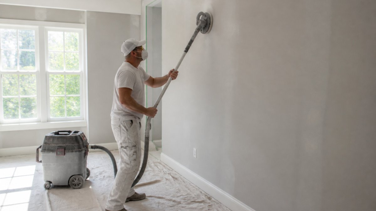 A professional painter in clean work clothes sanding a wall with a sanding pole, fine dust visible in the air, canvas drop cloths covering the floor, and a shop vacuum standing nearby in a bright residential room