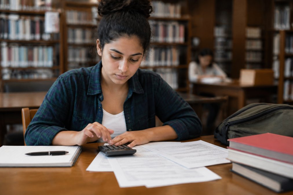A college student sitting at a library table with a laptop, calculator, and financial aid papers spread out in front of them
