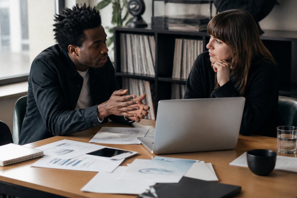 Independent music artist and label owner discussing streaming analytics and financial data on a laptop during a business meeting in a modern office, with charts and documents visible on desk